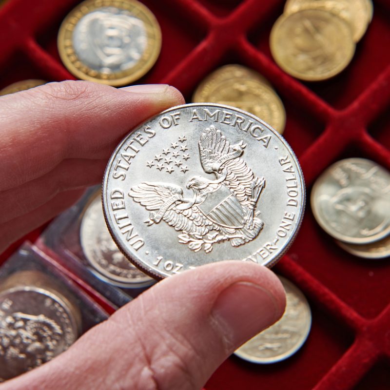 A hand holding a silver U.S. dollar coin with an eagle design, in front of a collector’s coin tray.