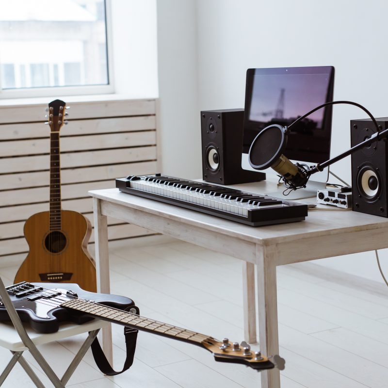 Home music studio setup with electric guitar, acoustic guitar, MIDI keyboard, speakers, microphone, and computer on a white wooden desk.