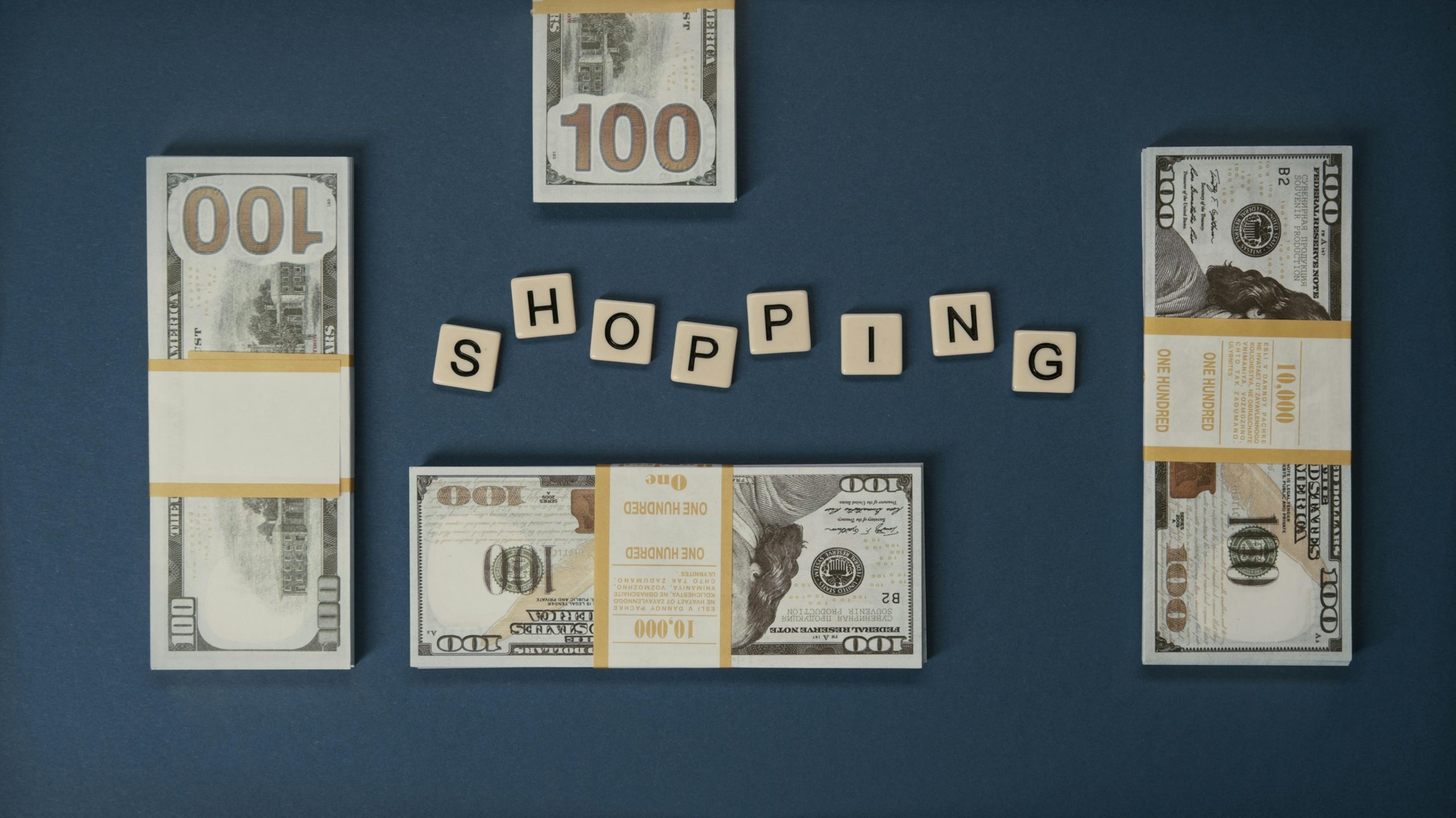 Bundles of hundred-dollar bills arranged around wooden tiles spelling “SHOPPING” on a dark background