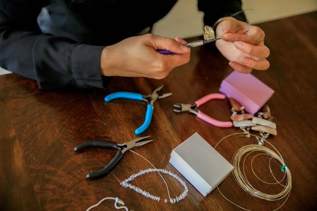 Hands using pliers to redesign and craft jewelry from pre-owned gold and gemstone pieces on a wooden table